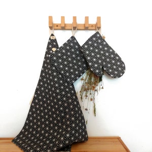 May include: A black and white geometric patterned oven mitt and matching kitchen towel hanging on a wooden rack. The pattern is a repeating design of white stars on a black background.