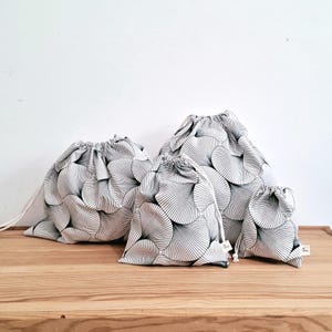 Può includere: Quattro sacchetti con coulisse di diverse dimensioni, disposti su una superficie di legno. I sacchetti sono bianchi con un motivo nero a forma di ventaglio. Ogni sacchetto ha un cordoncino bianco. Ideali per riporre oggetti.