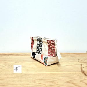 May include: A small, rectangular fabric pouch with a patchwork design. The pouch features vertical panels of cream, red, black, and floral patterns. The pouch is sitting on a wooden surface.
