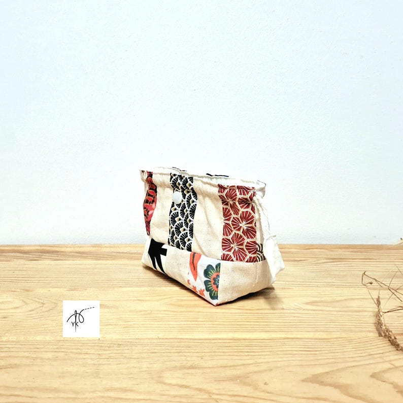 May include: A small, rectangular fabric pouch with a patchwork design. The pouch features vertical panels of cream, red, black, and floral patterns. The pouch is sitting on a wooden surface.