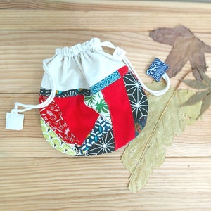 Pode incluir: Uma bolsa de cordão branco com um design de patchwork. A bolsa é feita de diferentes tecidos com padrões vermelhos, verdes, azuis e pretos. A bolsa está sobre uma superfície de madeira.