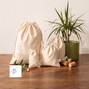 Könnte beinhalten: Ein Set aus vier cremefarbenen Baumwollbeuteln mit Kordelzug in verschiedenen Größen, jeweils mit einem kleinen Stoffetikett an der Seite. Die Beutel sind zusammen angeordnet, mit einem kleinen Logo, das eine Nadel und Faden sowie YK zeigt.
