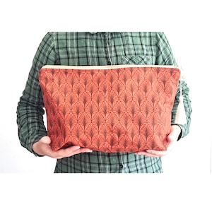 Pode incluir: Uma bolsa retangular com fecho de correr apresentando um padrão repetitivo em forma de leque com linhas castanho-escuras sobre um fundo laranja queimado. A bolsa tem um fecho claro na borda superior e uma pequena borla branca na lateral.