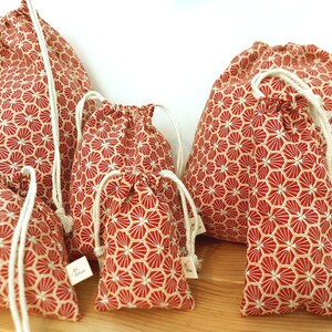 May include: Set of five red and white drawstring bags with a geometric pattern. The bags are made of fabric and have a white drawstring closure. The bags are arranged in a row from largest to smallest.