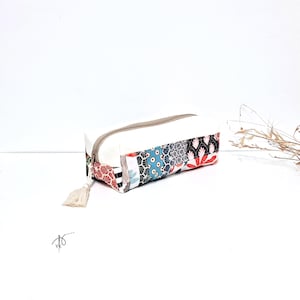 May include: Rectangular pencil case with a white body and a patterned panel featuring red, black, blue, and grey floral and geometric designs. The case has a beige zip and a tassel. The case is on a white surface.