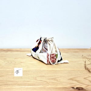 May include: A small drawstring bag made from patchwork fabric. The bag features a mix of patterns, including floral and geometric designs, in shades of red, blue, and cream. The bag is sitting on a wooden surface.