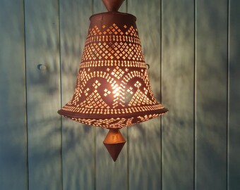 May include: A hanging, ornate, bronze-colored metal lamp with a bell-shaped shade. The shade features a detailed, geometric pattern of cut-out designs that allow light to shine through. The lamp is suspended from a chain and has a pointed finial at the bottom.