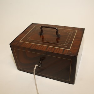 May include: A vintage wooden-look metal money box with a key and string attached. The box has a decorative inlay and a metal handle.
