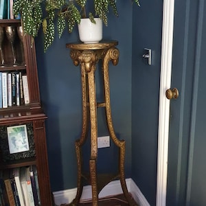 May include: A gold-colored, ornate plant stand with a white potted plant featuring green leaves with white spots. The stand has a round top and a detailed, carved design. The background is a dark blue wall.