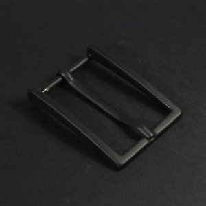 以下が含まれることがあります： シンプルな長方形のデザインの黒い金属製のベルトバックルです。