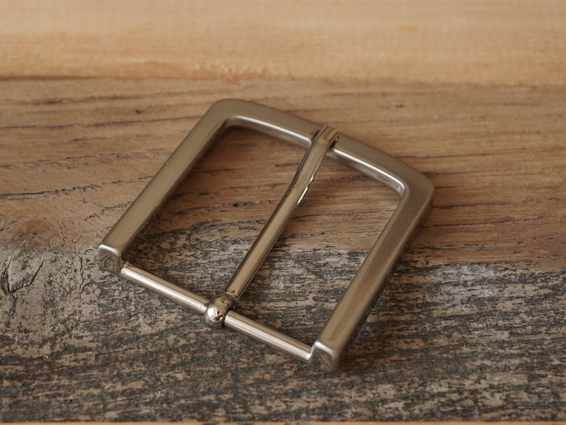 May include: A silver metal belt buckle with a rectangular shape and a smooth finish.