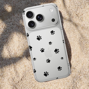 May include: Clear iPhone case with black paw prints scattered across the back. The case has a transparent design, allowing the phone's color to show through. The phone case is lying on a sandy surface.