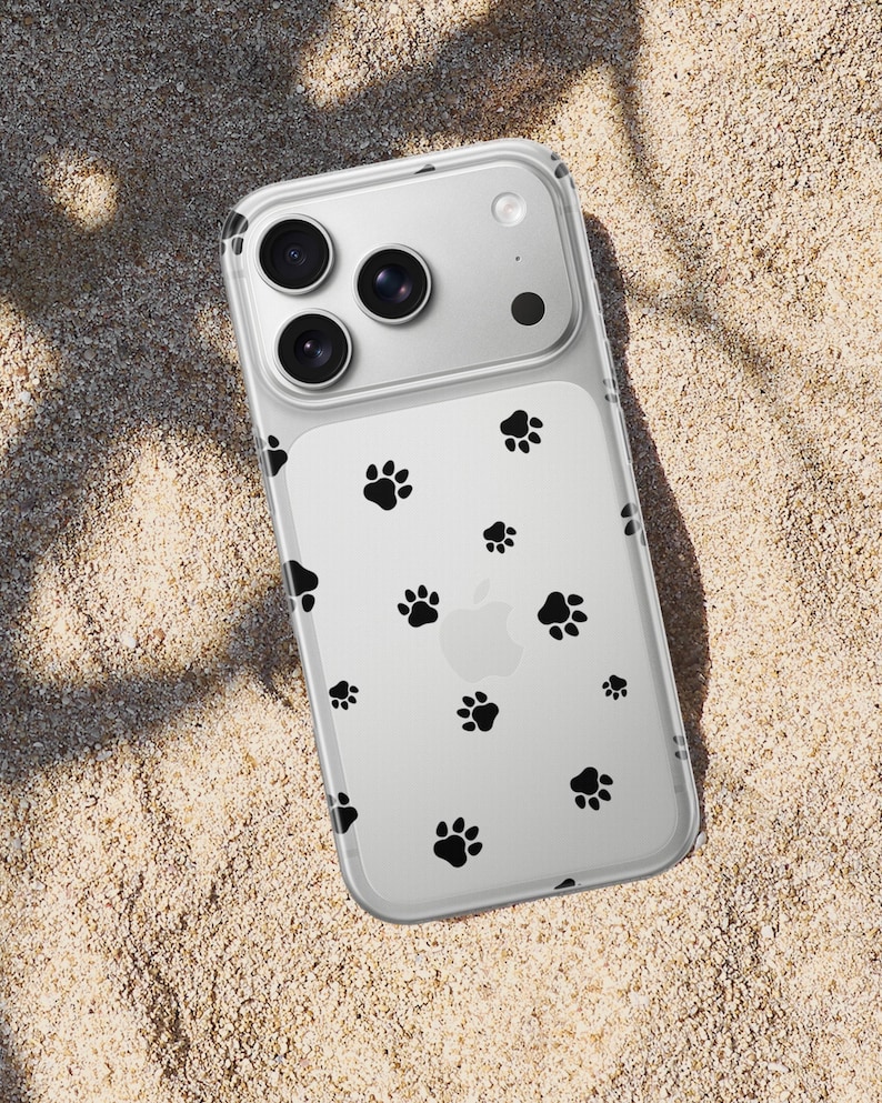 May include: Clear iPhone case with black paw prints scattered across the back. The case has a transparent design, allowing the phone's color to show through. The phone case is lying on a sandy surface.