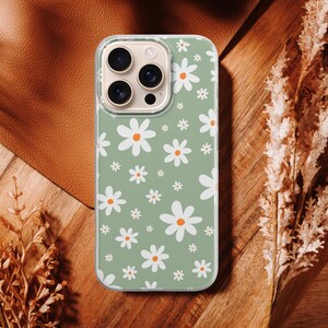 Pode incluir: Uma capa de telefone com um padr&atilde;o de margaridas. A capa tem um fundo verde claro com margaridas brancas e centros laranja. A capa do telefone est&aacute; em uma superf&iacute;cie de madeira com plantas secas.