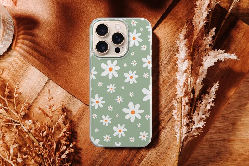 Pode incluir: Uma capa de telefone com um padr&atilde;o de margaridas. A capa tem um fundo verde claro com margaridas brancas e centros laranja. A capa do telefone est&aacute; em uma superf&iacute;cie de madeira com plantas secas.