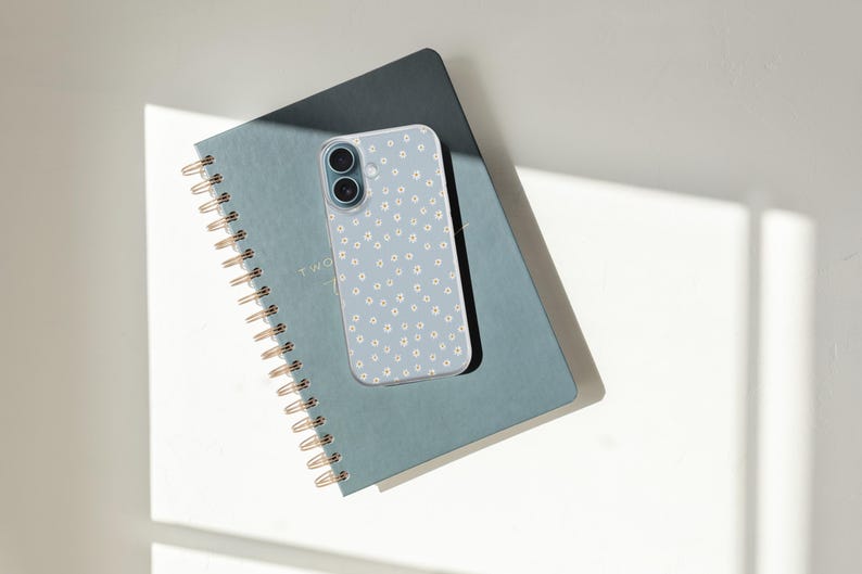 Pode incluir: Uma capa de telefone azul claro com um padr&atilde;o de margaridas brancas repousa sobre um caderno azul correspondente. O caderno tem uma encaderna&ccedil;&atilde;o em espiral dourada e o texto "TWO" &eacute; vis&iacute;vel. A cena &eacute; iluminada por luz natural.
