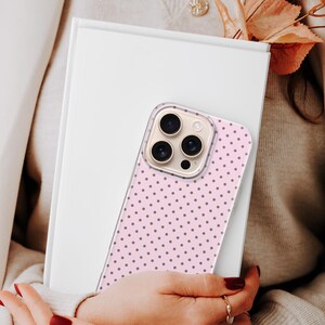 May include: A pink phone case with a polka dot pattern is held over a white book. The phone case has a clear border and a series of camera lenses. The background includes a person wearing a sweater and gold jewelry.
