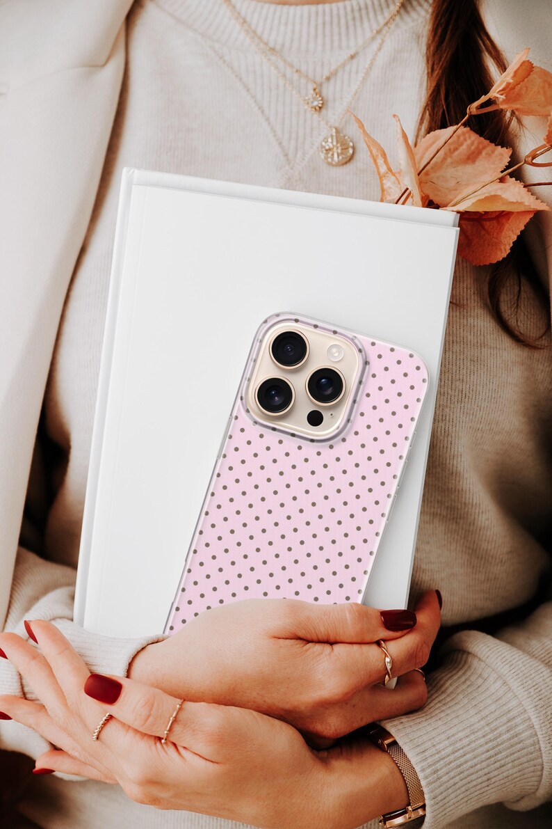 May include: A pink phone case with a polka dot pattern is held over a white book. The phone case has a clear border and a series of camera lenses. The background includes a person wearing a sweater and gold jewelry.
