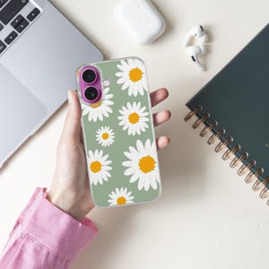 Pode incluir: Capa de telefone transparente com um padr&atilde;o de margaridas com p&eacute;talas brancas e centros amarelos em um fundo verde s&aacute;lvia. A capa &eacute; segurada perto de um laptop, um caderno e fones de ouvido sem fio.