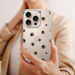 May include: Clear phone case with a pattern of black paw prints. The case is held up in front of a person. The phone has a triple-lens camera.