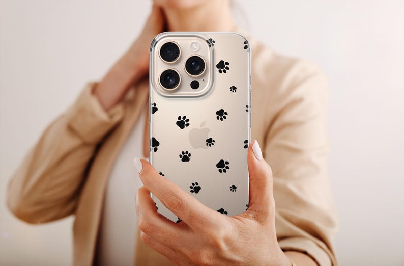 May include: Clear phone case with a pattern of black paw prints. The case is held up in front of a person. The phone has a triple-lens camera.