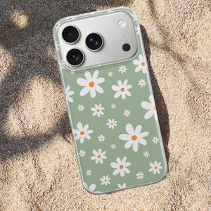 Pode incluir: Capa de telefone transparente com um padr&atilde;o de margaridas brancas com centros laranja sobre um fundo verde. A capa est&aacute; sobre uma superf&iacute;cie arenosa.