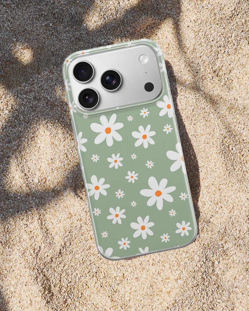 Pode incluir: Capa de telefone transparente com um padr&atilde;o de margaridas brancas com centros laranja sobre um fundo verde. A capa est&aacute; sobre uma superf&iacute;cie arenosa.