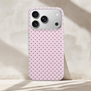 May include: A pink phone case with a repeating pattern of small, brown polka dots. The case has a clear border and a silver camera module. The phone case is leaning against a neutral-colored wall.