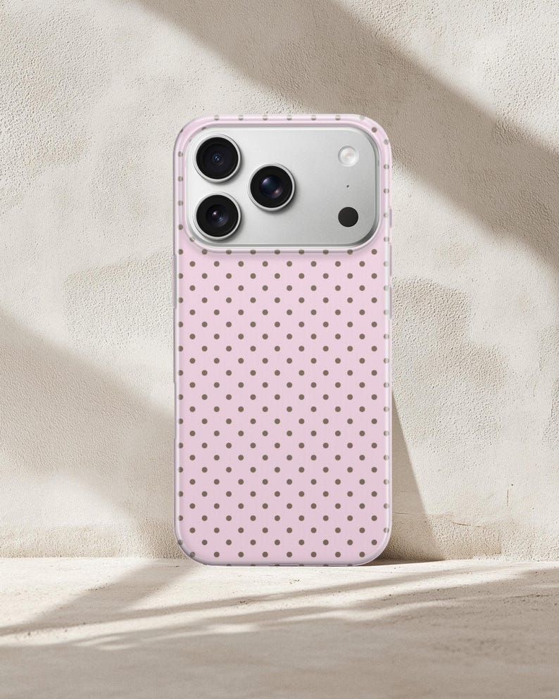 May include: A pink phone case with a repeating pattern of small, brown polka dots. The case has a clear border and a silver camera module. The phone case is leaning against a neutral-colored wall.