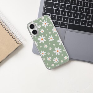 Pode incluir: Uma capa de telefone verde com um padr&atilde;o de margaridas brancas est&aacute; sobre um laptop. Um caderno espiralado est&aacute; &agrave; esquerda. A capa tem uma borda transparente e uma c&acirc;mera de lente dupla. O teclado do laptop &eacute; preto.