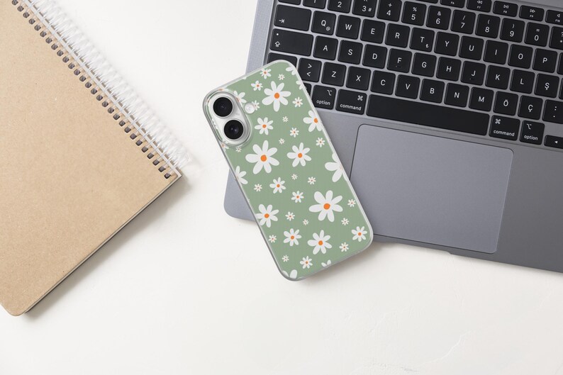Pode incluir: Uma capa de telefone verde com um padr&atilde;o de margaridas brancas est&aacute; sobre um laptop. Um caderno espiralado est&aacute; &agrave; esquerda. A capa tem uma borda transparente e uma c&acirc;mera de lente dupla. O teclado do laptop &eacute; preto.