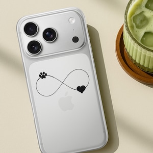 May include: Clear phone case with a black infinity symbol design featuring a paw print and heart. The case is on a white phone with a triple-lens camera. A glass of green drink sits on a wooden coaster.