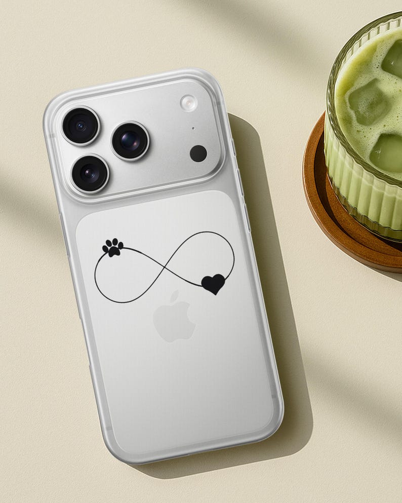 May include: Clear phone case with a black infinity symbol design featuring a paw print and heart. The case is on a white phone with a triple-lens camera. A glass of green drink sits on a wooden coaster.