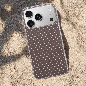 May include: A smartphone case with a brown background and a pattern of small, evenly spaced pink polka dots. The case has a clear border and a camera module with three lenses and a flash.