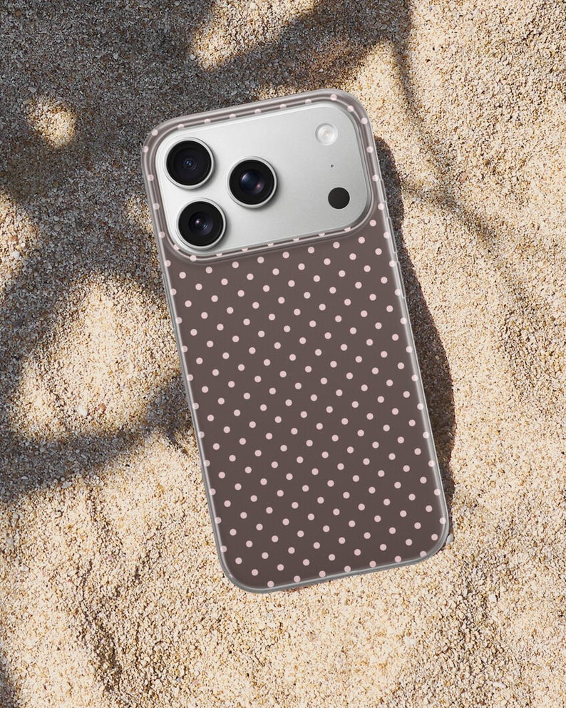 May include: A smartphone case with a brown background and a pattern of small, evenly spaced pink polka dots. The case has a clear border and a camera module with three lenses and a flash.