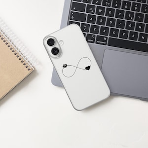 May include: A white phone case featuring a black infinity symbol with a paw print and heart design. The phone case is on a laptop, next to a spiral-bound notebook. The laptop keyboard is visible.