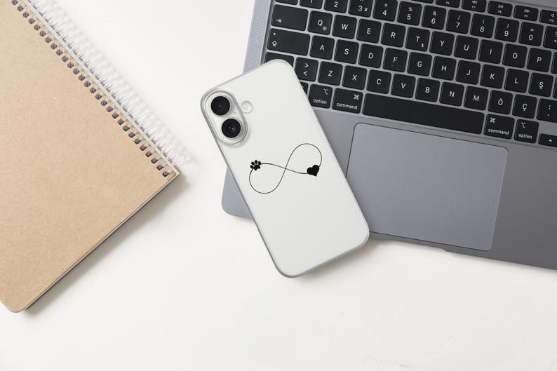 May include: A white phone case featuring a black infinity symbol with a paw print and heart design. The phone case is on a laptop, next to a spiral-bound notebook. The laptop keyboard is visible.