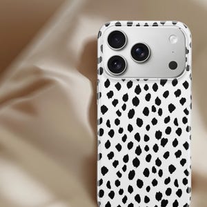 May include: A white phone case with a black dalmatian spot pattern. The case has a clear border and a camera cutout with three lenses and a flash. The phone case is designed to protect the phone.