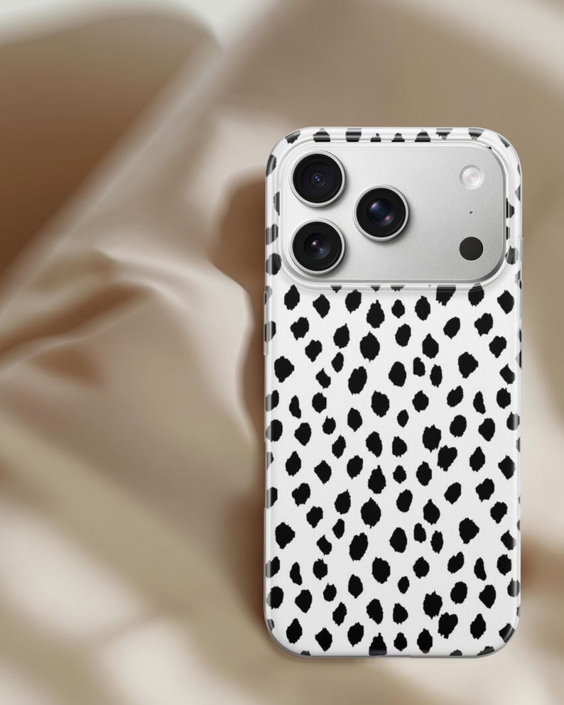 May include: A white phone case with a black dalmatian spot pattern. The case has a clear border and a camera cutout with three lenses and a flash. The phone case is designed to protect the phone.
