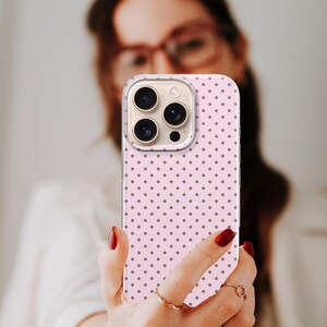 May include: A pink phone case with a repeating pattern of small, dark gray polka dots. The phone case has a clear border and is held up in front of a person. The phone case has a camera with three lenses.