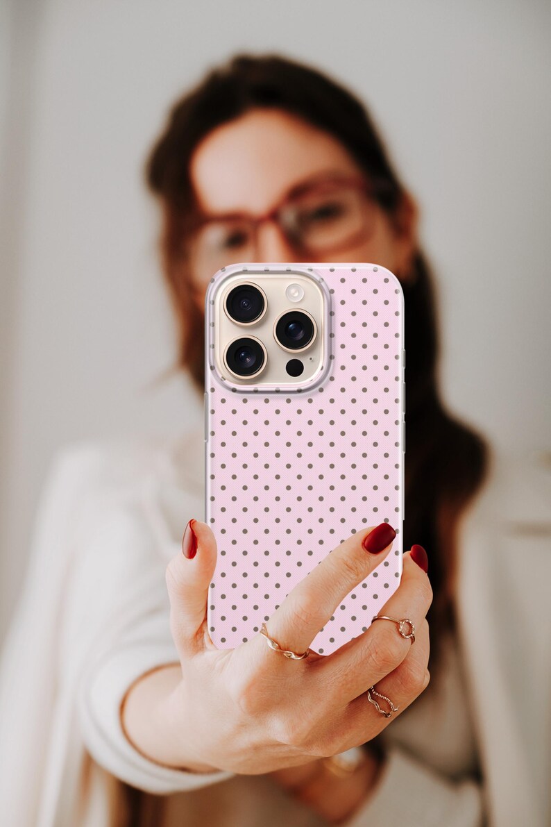 May include: A pink phone case with a repeating pattern of small, dark gray polka dots. The phone case has a clear border and is held up in front of a person. The phone case has a camera with three lenses.