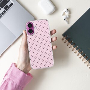 May include: A pink phone case with a polka dot pattern is held in a person's hand. The phone case is next to a laptop, a dark green spiral-bound notebook, and wireless earbuds. The notebook has gold lettering that reads "TWO THOUSAND twenty-two".