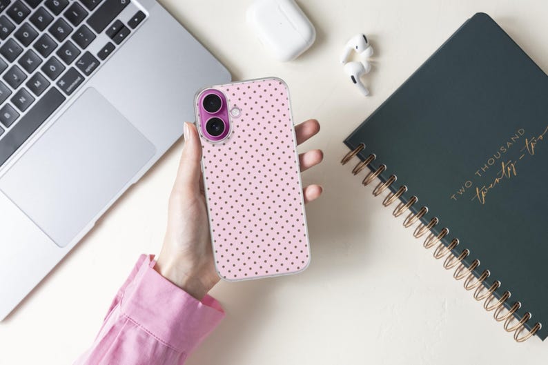 May include: A pink phone case with a polka dot pattern is held in a person's hand. The phone case is next to a laptop, a dark green spiral-bound notebook, and wireless earbuds. The notebook has gold lettering that reads "TWO THOUSAND twenty-two".