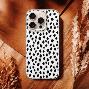 May include: A white phone case with a black dalmatian spot pattern. The case has a clear border and a camera cutout. The phone case is on a wooden surface with dried plants and a leather background.