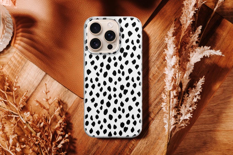 May include: A white phone case with a black dalmatian spot pattern. The case has a clear border and a camera cutout. The phone case is on a wooden surface with dried plants and a leather background.