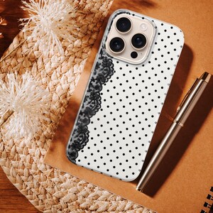 May include: A white phone case with black polka dots and a black lace design on the side. A silver pen and a brown notebook are next to the phone case. The phone case is on a woven surface with dried flowers.