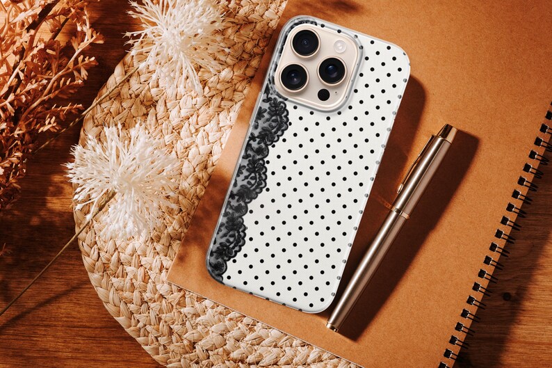 May include: A white phone case with black polka dots and a black lace design on the side. A silver pen and a brown notebook are next to the phone case. The phone case is on a woven surface with dried flowers.