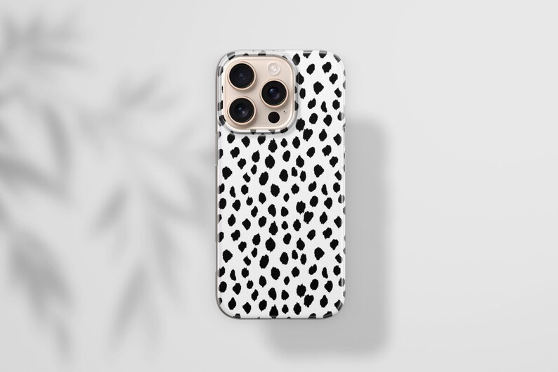 May include: A white phone case with a black dalmatian spot pattern. The case has a clear border and a camera module with multiple lenses. The background is a light gray surface with a shadow of leaves.