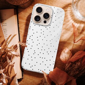 May include: A white phone case with a black polka dot pattern. The phone case is on a wooden surface, next to a book, dried flowers, and a glass of water. The case has a clear border.
