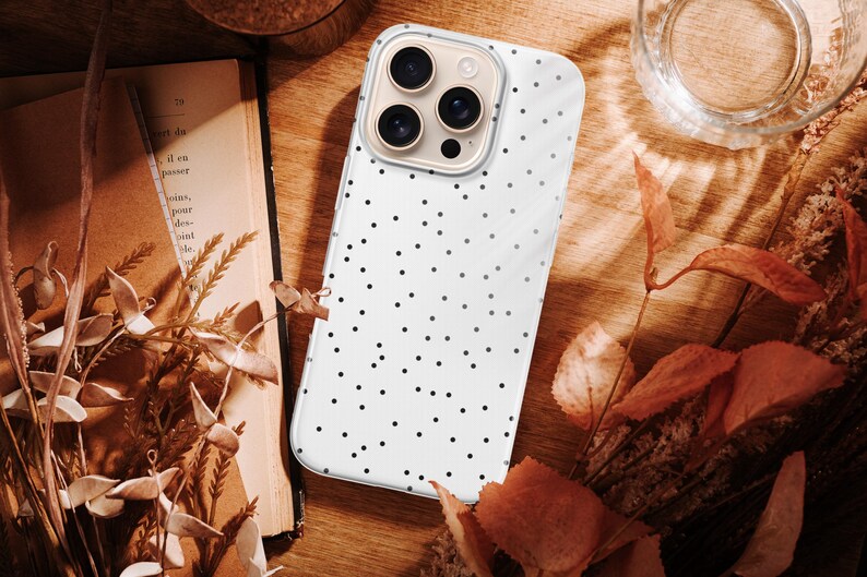 May include: A white phone case with a black polka dot pattern. The phone case is on a wooden surface, next to a book, dried flowers, and a glass of water. The case has a clear border.